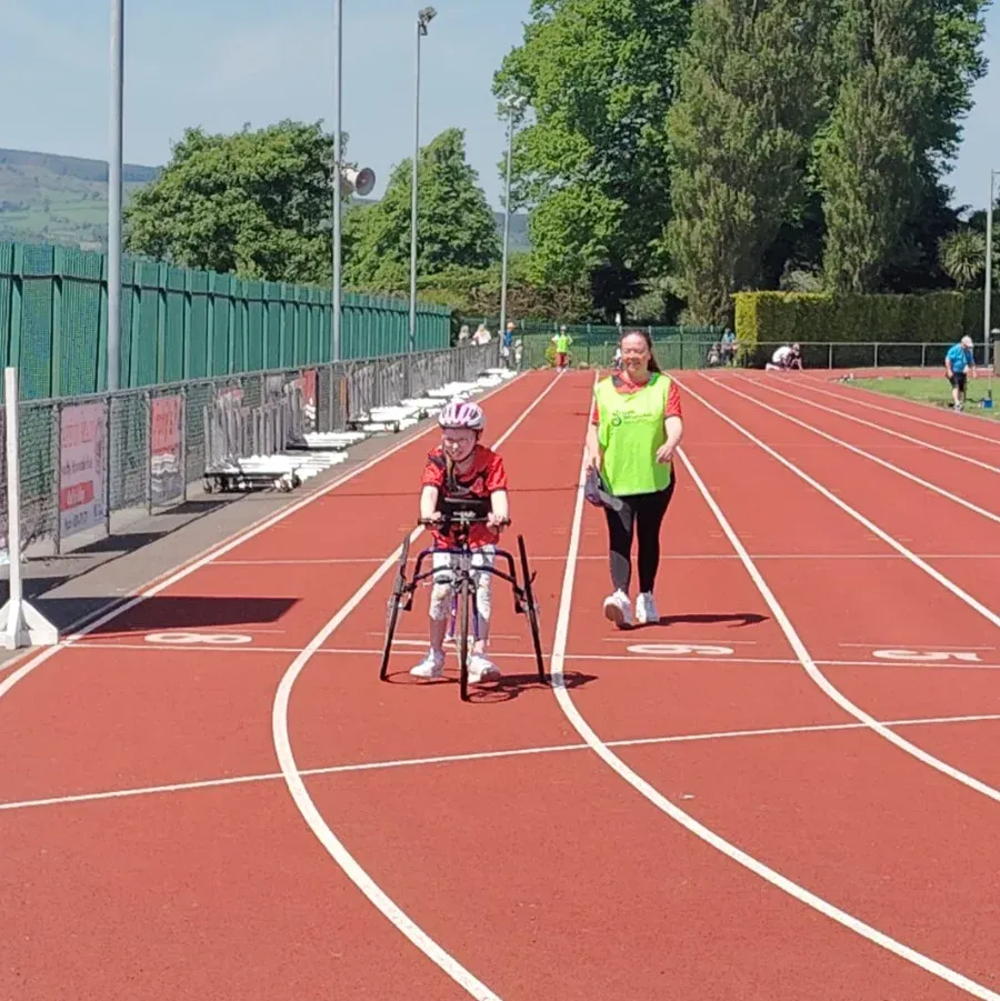 junior on a para athletics bike in a running track accompanied by a volunteer